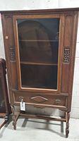 Full front view of the antique wooden curio cabinet showing glass door with carved wood detailing and drawer below.
