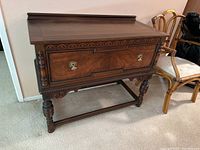 Front view of antique walnut server showing carved top trim, drawer with metal ring pulls, and turned legs with stretchers.