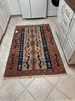 Photo of a small rectangular area rug with geometric motif in the kitchen on tile floor. Lace-like fringe tassels on one end of the rug.