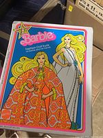 Photo of Barbie Fashion Doll Trunk box with vibrant 70s style illustration of Barbie in a flowing orange patterned dress and another Barbie in a white embellished dress for her friends. The box is closed.
