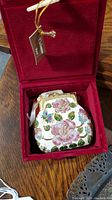 Decorative miniature floral enamel bag with gold-tone details, housed in a red velvet box, photographed on wooden table.