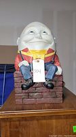 Front view of Humpty Dumpty statue, showing the full figure seated on the brick wall base with an attached tag labeled 354, placed on a wooden surface.
