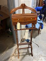 Front view of the mid-century wooden valet stand showing the hanger, two drawers with brass knobs, trouser bar, and four legs in a warehouse setting.