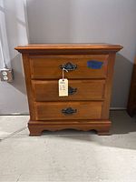 Front view of wooden nightstand table showing three drawers with decorative metal handles, curved bottom edge, and natural wood finish.