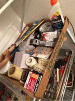 View of assorted painting supplies in cardboard box including glue bottle, paint bottles, rollers, tape and various accessories.