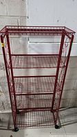 Full front view of the red metal rolling shelf showing five wire mesh shelves and caster wheels.