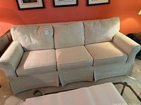 Full view of a beige fabric three-seater sofa with three seat cushions and three back cushions, rolled arms, and skirted base, placed against an orange wall below framed pictures.