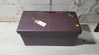 Full view of brown collapsible storage ottoman with padded lid on concrete floor with wall background.