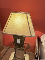 Table lamp shown fully assembled on a glass table against a coral wall with beige hexagonal fabric lampshade and dark metal base.