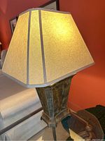 Photo showing close-up side view of the table lamp, highlighting the trapezoidal beige fabric shade with brown trim and dark distressed metal base.
