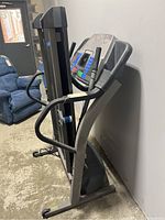 Side view of gray treadmill showing digital display and side handles against a wall with a blue recliner chair in the background.