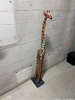 Full view of a tall wooden giraffe statue painted brown with beige and dark spots, standing against a concrete wall on a textured rectangular black base.
