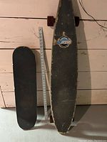 Photo of two skateboards standing upright against a wall with a measuring stick showing size comparison. One is a longboard and the other a shortboard.