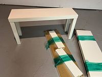 One assembled white Ikea Lack bench next to two flat packed benches wrapped in green tape on concrete floor.