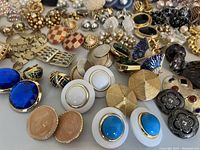 Photo showing a variety of clip-on earrings with blue faceted stones, white enamel beads, gold-tone textured discs, and checkered red and beige pieces arranged close-up.