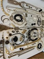 Overview of assorted costume jewelry including several chain necklaces, bracelets, stud and dangle earrings, and brooches with black beads and rhinestones laid out on white surface.