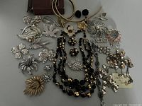 Photo of the full lot showing vintage silver-tone brooches mostly floral and leaf designs, black glass multi-strand necklace with matching earrings centrally placed, boxed Avon belt with floral charms, and two Stella & Dot necklaces with charms. One brooch missing a stone visible.