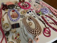 Wide view of multi-strand necklaces, beaded bracelets, earrings, and brooches arranged on white surface, showing various costume jewelry items in pink and purple tones.