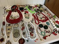 Overview of costume jewelry lot showing red, green, and white necklaces, bracelets, earrings, brooches, and other pieces spread on a white surface.