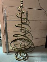 Full view of the vintage metal spiral Christmas tree ornament display stand against a wooden wall and concrete floor, with a measuring stick for scale showing approximately 39 inches tall.