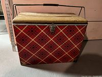 Front view of the rectangular red and black plaid metal camping cooler with a metal handle and latch, showing size reference with a nearby ruler.