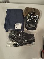 Photo of a folded blue pillowcase, a brown distressed baseball cap, and black gloves in sealed packaging arranged on a flat surface.