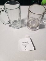 Two glass mugs side by side on a white surface with auction tag number 3 in front.
