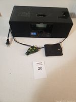 Front view of the JVC black audio device with attached power cord, dock connector on top, control knob, digital display, with small soft pouch and toy motorcycle in front on a table.
