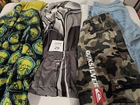 Photo of Grinich sweater with yellow and green abstract faces on black background, bike shirt, camo shorts, and jeans