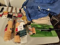 Photo showing paint brushes, foam rollers, a green paint tray with roller frame, and folded protective aprons or coveralls all laid out on a blue sheet.