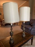 Two mid-century modern metal base table lamps with tall off-white cylindrical fabric shades, positioned on a table in a living room setting under natural light.