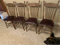 Four walnut kitchen chairs with carved backs and leather seats arranged in a row on carpeted floor.