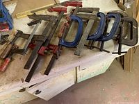 5 C-clamps and 4 bar clamps on a workbench, showing metal construction with screw handles and wear from use