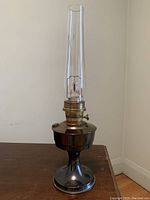 Full view of the vintage Aladdin brass oil lamp standing on a wooden surface against a plain wall. Shows the complete lamp with glass chimney.