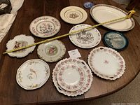 All plates and platters arranged on a wooden table including plates with portraits, floral patterns, and blue scenic designs