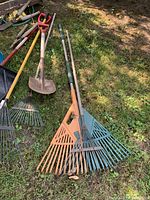 Photo showing several leaf rakes with wooden handles and plastic tines, as well as shovels and other garden tools laid on grass.