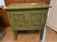 Front view of green wooden cabinet with hand-carved floral panels on doors. One metal knob on right door missing.