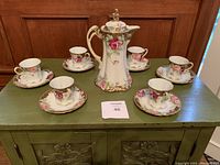 Full set showing 6 tea cups and saucers arranged around the tea pot on a green table in front of a wooden door.
