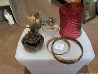 Lot displayed on white plastic storage bin, showing all components including glass oil reservoir, brass burner, cranberry glass shade, and brass decorative metal pieces.