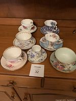 Seven assorted vintage porcelain teacups with matching saucers arranged on wooden cabinet surface
