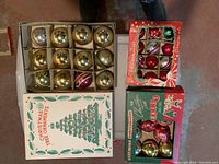 Photo showing four boxes of mid-century Christmas ornaments including a white box with green tree design filled with gold and silver glass balls and three smaller red and green boxes with assorted colorful baubles