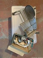 Photo shows metal manual meat slicer with round blade and ridged metal platform for slicing, placed on a plastic storage container.