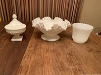 Set of three white milk glass items on wooden table: covered candy dish, ruffled bowl, hobnail vase.