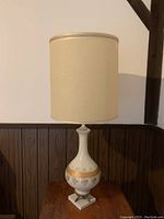 Porcelain lamp standing on a wooden surface with beige cylinder shade unlit, showing full lamp profile and design details.