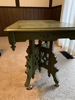 Side view showing the table's carved and cut-out leg design, metal castors, and some paint damage on the top surface