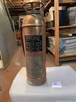 Full view of the antique copper fire extinguisher showing its height and cylindrical shape with handle and top ring, standing on a white plastic bin in a basement setting.