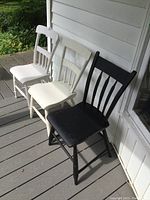 Three wooden painted chairs on porch, two white and one black in color.