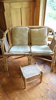 Two-seat rattan bench with beige cushions and small rattan stool on wooden floor.