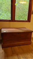 Photo showing the front side of the large vintage wooden blanket box under a window, chest is rectangular with a slightly raised base molding and hinged lid, surface is worn with scratches.
