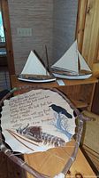 Two wooden model boats with white sails on a wooden shelf and indigenous leather art canvas with feathers below them.